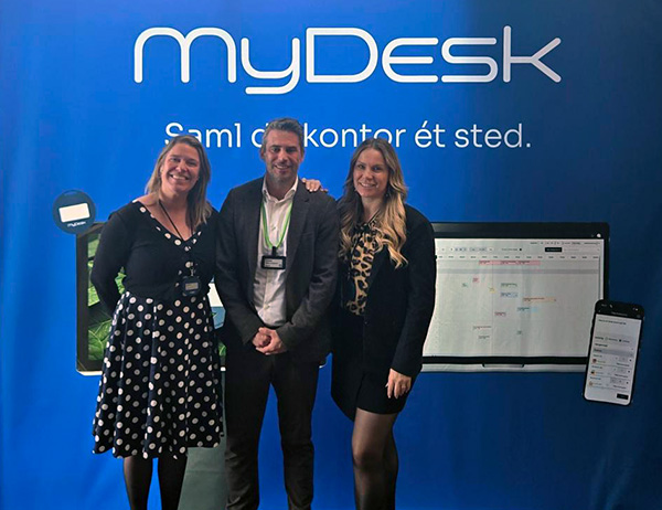 Three smiling professionals pose in front of a blue myDesk booth backdrop at a trade show, with a large monitor and phone graphic in view.
