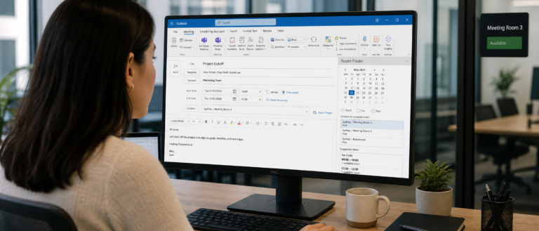 Person viewed from behind at a desk, monitor shows Outlook with an email/meeting form and room finder panel.