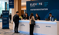 Visitors register at a conference registration desk labeled Elevate 2025 in a blue booth area with staff assisting attendees.