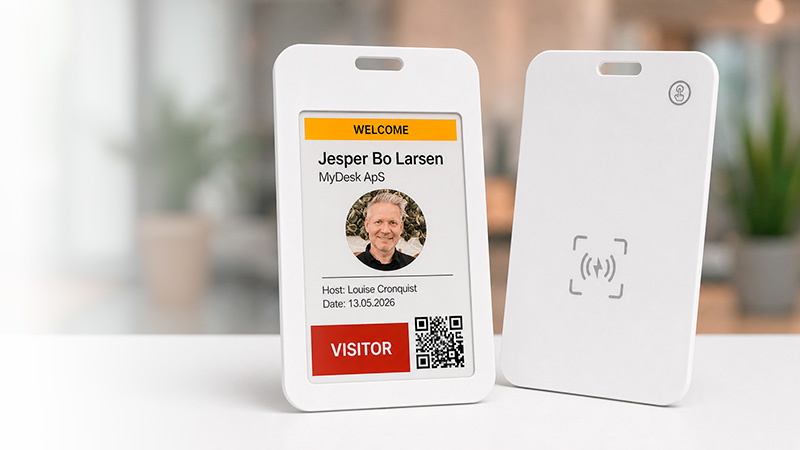 Two white visitor badges on a desk; the front badge shows name Jesper Bo Larsen, host Louise Cronquist, a photo, a QR code, and a red VISITOR label with a yellow header. Back badge shows a contactless area icon.